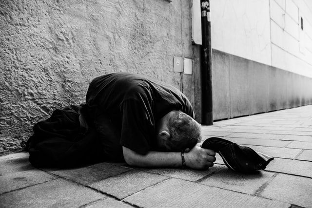 A greyscale image of a man kneeling on a sidewalk, cap in hand, forehead pressed to the ground.
